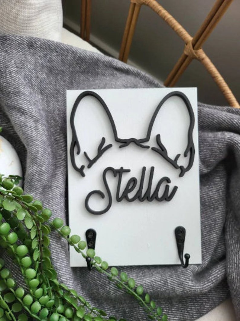 May include: White wooden key holder with a black silhouette of a French bulldog's head and the name "Stella" in black lettering. Two black hooks are attached to the bottom of the holder.