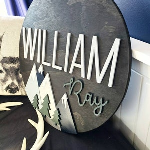 May include: A round wooden sign with a dark wood stain. The sign features a mountain scene with white, gray, and blue mountains, green trees, and the names "William Ray" in white and black lettering.