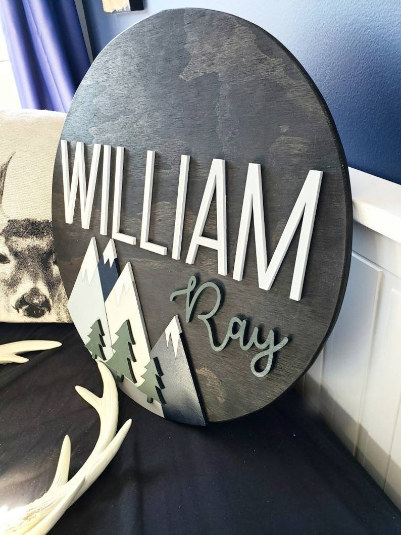 May include: A round wooden sign with a dark wood stain. The sign features a mountain scene with white, gray, and blue mountains, green trees, and the names "William Ray" in white and black lettering.