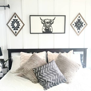 May include: A black and white silhouette of a Highland cow's head is framed and hung on a white wall above a black headboard with a white bedspread and several throw pillows. Two diamond-shaped wall decorations with black and white geometric patterns are also hung on the wall.