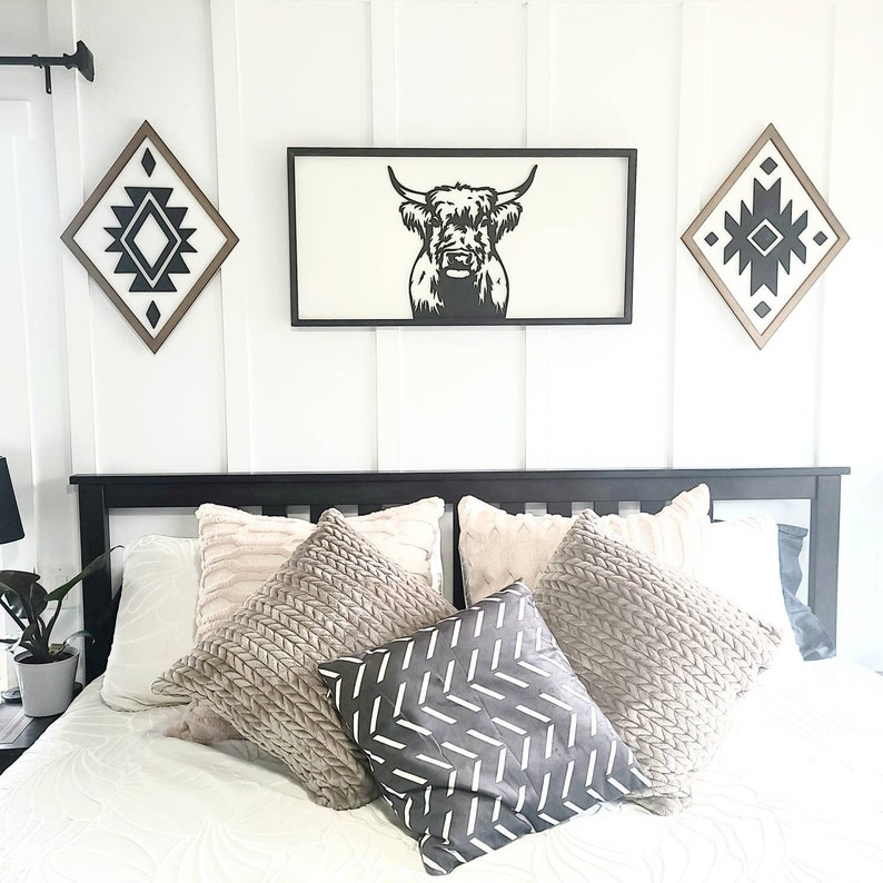 May include: A black and white silhouette of a Highland cow's head is framed and hung on a white wall above a black headboard with a white bedspread and several throw pillows. Two diamond-shaped wall decorations with black and white geometric patterns are also hung on the wall.