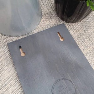 May include: A gray wooden sign with two holes for hanging. The sign has a distressed finish and the text "The Little  Treasury" is printed in a circle in the lower right corner.
