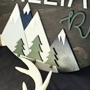 May include: A wooden sign with a mountain scene and the name "Raylan" in cursive script. The mountains are white, blue, and gray, and the trees are green. The sign is on a dark brown background.