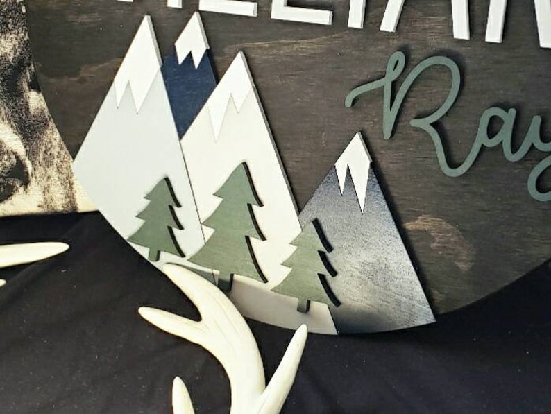 May include: A wooden sign with a mountain scene and the name "Raylan" in cursive script. The mountains are white, blue, and gray, and the trees are green. The sign is on a dark brown background.