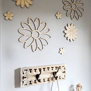 K&ouml;nnte beinhalten: Ein wei&szlig;er Holz-Kleiderhaken mit geschnitzten Details und drei schwarzen Metallhaken. Der Haken ist an einer hellgrauen Wand mit mehreren h&ouml;lzernen Wanddekorationen in Form von G&auml;nsebl&uuml;mchen befestigt.