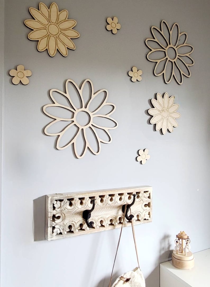 K&ouml;nnte beinhalten: Ein wei&szlig;er Holz-Kleiderhaken mit geschnitzten Details und drei schwarzen Metallhaken. Der Haken ist an einer hellgrauen Wand mit mehreren h&ouml;lzernen Wanddekorationen in Form von G&auml;nsebl&uuml;mchen befestigt.
