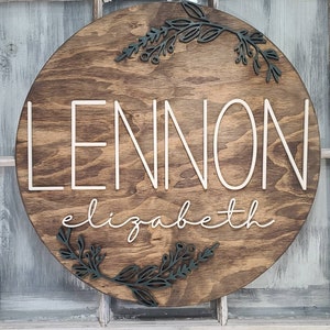 May include: A round wooden sign with the name "LENNON elizabeth" written in white script. The sign is decorated with dark green leafy branches.