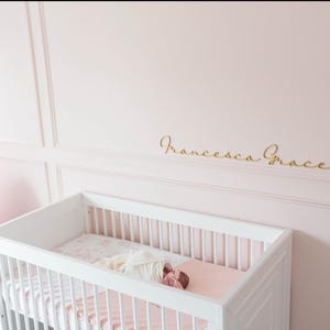 May include: A white crib with a pink mattress and bedding is in a pink room. The name "Francesca Grace" is written in gold script on the wall above the crib. A gold sconce with a white shade is on the wall.