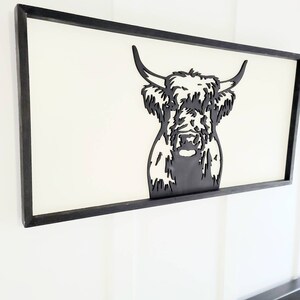May include: Black and white silhouette of a Highland cow head in a black wood frame. The cow has long, shaggy hair and a friendly expression.