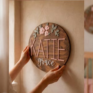May include: A circular wooden sign featuring the name "KATIE" and the word "grace" in pink, with pink flower and green leaf details. The sign is held against a wall. Text at the top reads "HANDMADE NURSERY DECOR" and at the bottom "FULLY CUSTOMIZABLE DESIGN".