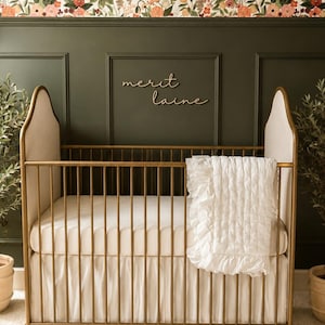 May include: A gold cot with a white fitted sheet, skirt, and a ruffled blanket. The cot is set against a dark green wall with the name "merit laine" in wooden letters. The wallpaper above the wall is floral.