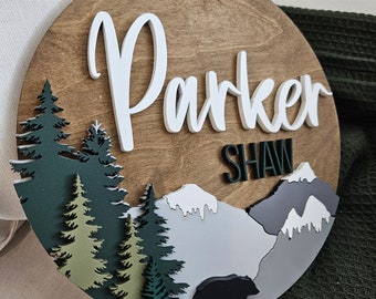 Puede incluir: Un letrero redondo de madera con una escena de montaña y un oso negro. El letrero está decorado con árboles verdes, montañas grises y nieve blanca. El nombre "Parker Shaw" está escrito en letras blancas en el letrero.