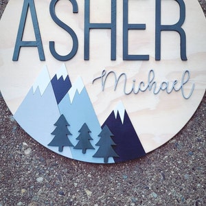 May include: A round wooden sign with the name "Asher Michael" written in black and gray lettering. The sign is decorated with blue and gray mountains and black trees.
