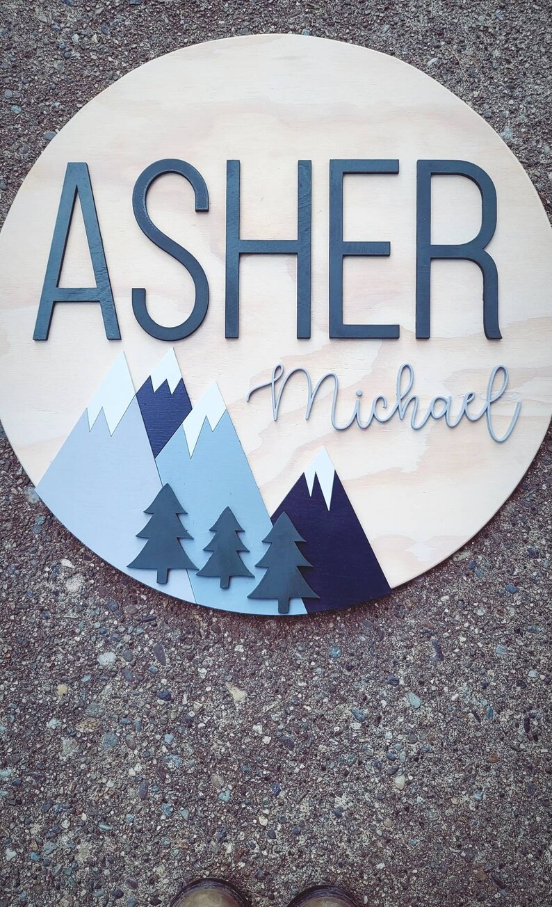 May include: A round wooden sign with the name "Asher Michael" written in black and gray lettering. The sign is decorated with blue and gray mountains and black trees.