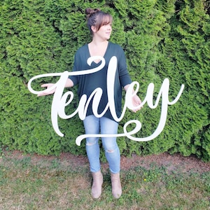 Peut inclure: Une pancarte en bois blanc avec le nom "Tenley" écrit en cursive. La pancarte est tenue par une personne portant une chemise vert foncé et un jean bleu.