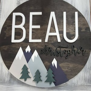 May include: A round wooden sign with the name "BEAU christopher" in white and black lettering. The sign features a mountain range with green pine trees and white snow peaks.