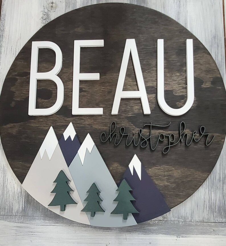 May include: A round wooden sign with the name "BEAU christopher" in white and black lettering. The sign features a mountain range with green pine trees and white snow peaks.