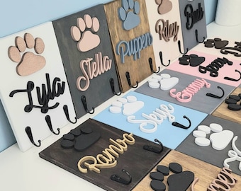 May include: A set of wooden dog leash hooks with paw prints and personalized names. The hooks are painted in various colors, including white, gray, pink, blue, and brown. The names on the hooks include Lyla, Stella, Puppen, Riley, Bud, Sammy, Skye, Emmy, Rambo, and Harper.