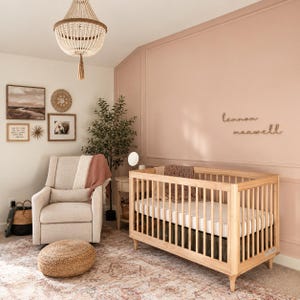 May include: A nursery room featuring a light wood cot with a white mattress, a beige upholstered recliner, a round woven jute ottoman, and a beaded chandelier. The wall is painted a soft dusty rose with wooden name cutouts reading lennon maxwell.
