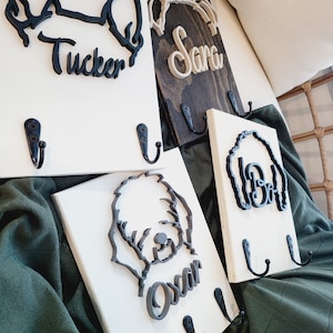 May include: Four white wooden dog-themed key hooks with black metal hooks. Each hook features a different dog breed silhouette and the name "Tucker", "Sara", "Oscar", and "Bella" in black lettering.