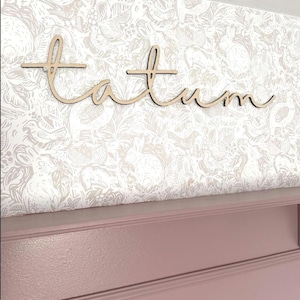 May include: A wooden sign with the name "Tatum" written in cursive script. The sign is mounted on a white wall with a floral pattern.
