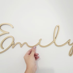 May include: A gold-colored wooden sign that spells out the name "Emily" in a cursive font. The sign is held by a hand against a white background. The sign is a decorative item.