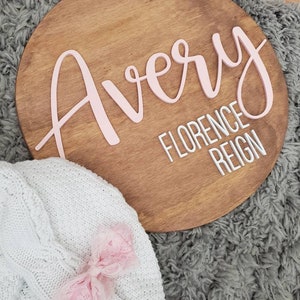 May include: A round wooden sign with the name "Avery" written in pink script. Below the name, the text "Florence Reign" is written in white.