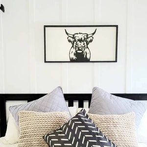 May include: Black and white silhouette of a Highland cow head in a black frame hanging above a bed with a white duvet and several pillows. The pillows are beige, gray, and black with a geometric pattern.