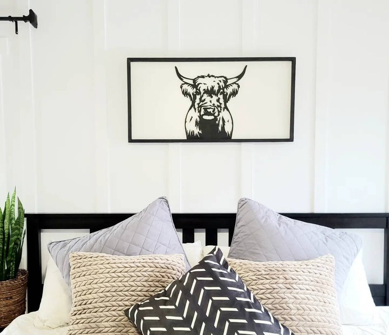 May include: Black and white silhouette of a Highland cow head in a black frame hanging above a bed with a white duvet and several pillows. The pillows are beige, gray, and black with a geometric pattern.