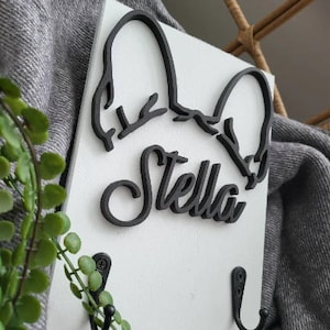 May include: White wooden wall mounted coat rack with two black hooks. The rack features a black silhouette of a dog's head with the name "Stella" written in a stylized font.