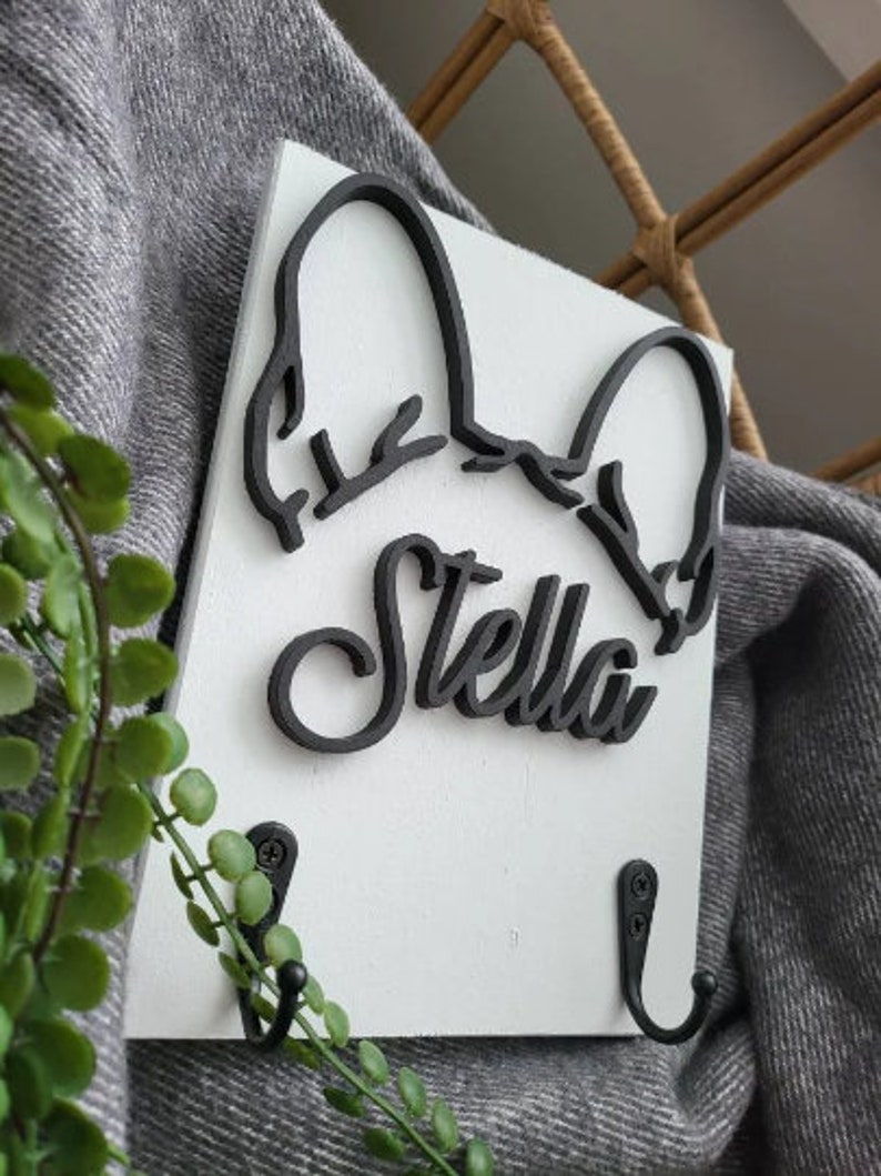 May include: White wooden wall mounted coat rack with two black hooks. The rack features a black silhouette of a dog's head with the name "Stella" written in a stylized font.