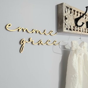 May include: A white wooden sign with the names "emmie grace" written in gold cursive script. The sign is hanging on a wall with a decorative wooden hook.