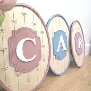 May include: Three oval wooden wall decorations spelling "CAR". Each has a different colour frame and centre. The "C" is in a pink frame, the "A" in a blue frame, and the "R" in a lavender frame. The background is a light wood grain with bow and flower patterns.