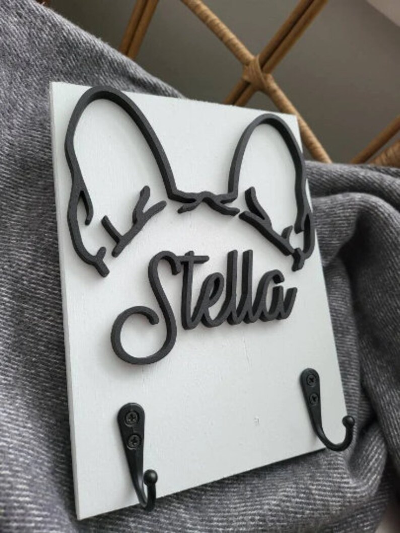 May include: White wooden key holder with a black silhouette of a French Bulldog's head and the name "Stella" in a cursive font. The key holder has two black metal hooks.