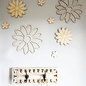 K&ouml;nnte beinhalten: Ein wei&szlig;er Holz-Kleiderst&auml;nder mit drei Haken und einem verzierten Design. Die Wand hinter dem Kleiderst&auml;nder ist mit Holz-G&auml;nsebl&uuml;mchenformen in verschiedenen Gr&ouml;&szlig;en dekoriert.