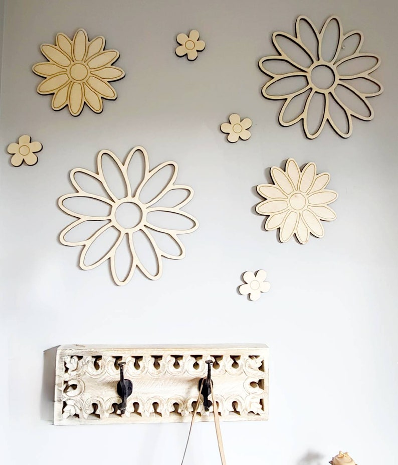 K&ouml;nnte beinhalten: Ein wei&szlig;er Holz-Kleiderst&auml;nder mit drei Haken und einem verzierten Design. Die Wand hinter dem Kleiderst&auml;nder ist mit Holz-G&auml;nsebl&uuml;mchenformen in verschiedenen Gr&ouml;&szlig;en dekoriert.