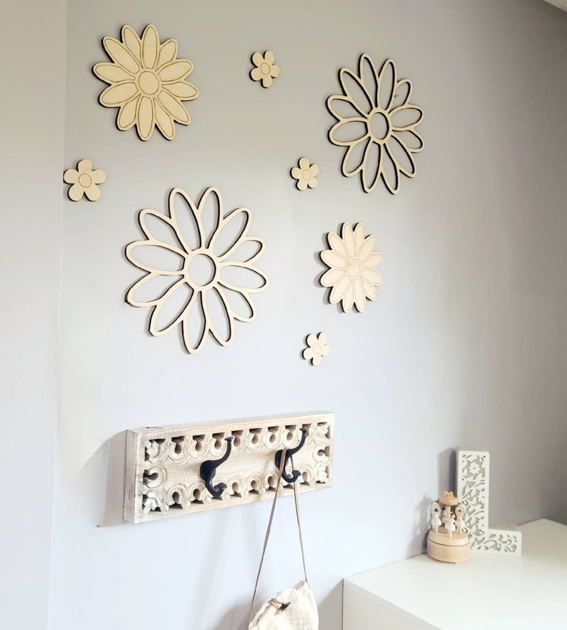 K&ouml;nnte beinhalten: Eine wei&szlig;e Wand mit einem Holz-Kleiderhaken mit zwei Haken. Die Wand ist mit mehreren Holz-Wanddekorationen in Form von G&auml;nsebl&uuml;mchen in verschiedenen Gr&ouml;&szlig;en verziert.