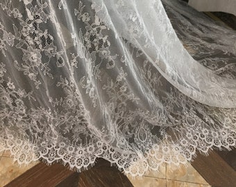 May include: A white lace fabric featuring an intricate floral pattern with a scalloped edge. The delicate material is sheer and lightweight, showcasing a detailed mesh background with embroidered botanical motifs throughout the entire length.