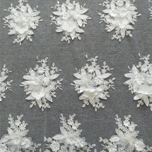 May include: White floral lace fabric with 3D flowers. The fabric has a delicate, intricate design with raised flowers and leaves. The fabric is perfect for wedding dresses, bridal accessories, and other special occasion garments.