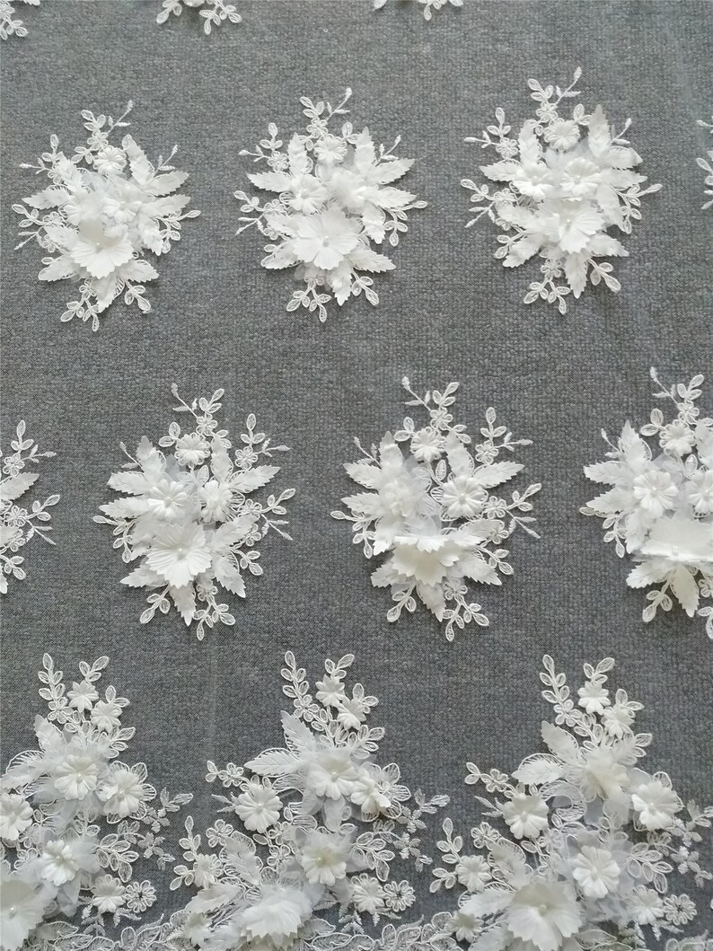May include: White floral lace fabric with 3D flowers. The fabric has a delicate, intricate design with raised flowers and leaves. The fabric is perfect for wedding dresses, bridal accessories, and other special occasion garments.