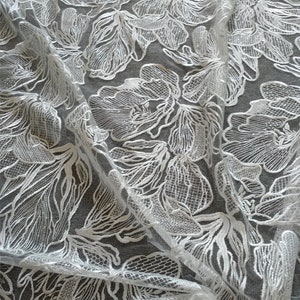 May include: White floral lace fabric with a delicate, intricate pattern. The lace is sheer and has a soft, flowing texture.