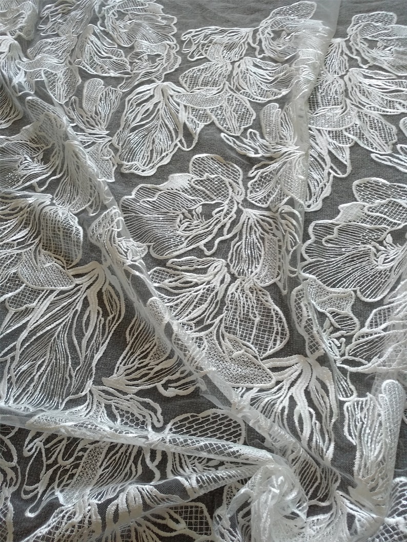 May include: White floral lace fabric with a delicate, intricate pattern. The lace is sheer and has a soft, flowing texture.