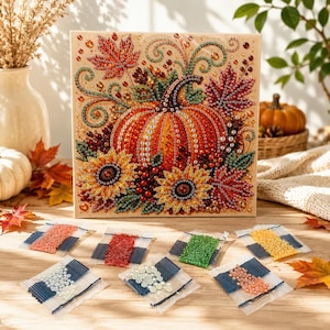 Pode incluir: Um kit de pintura de diamante quadrado com uma abóbora laranja central rodeada por girassóis amarelos, folhas de bordo vermelhas e vinhas verdes. A tela exibe seções numeradas e oito pequenos pacotes de contas coloridas.