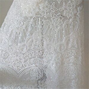 May include: White beaded lace fabric with a delicate floral pattern. The fabric is sheer and has a subtle sparkle.
