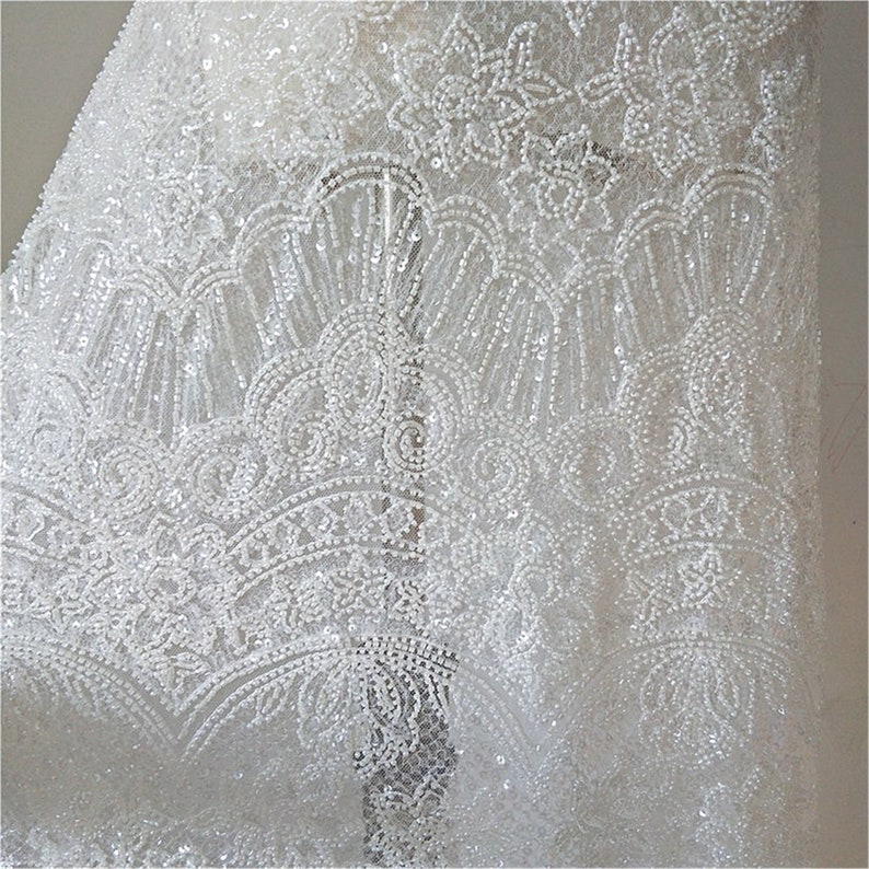 May include: White beaded lace fabric with a delicate floral pattern. The fabric is sheer and has a subtle sparkle.