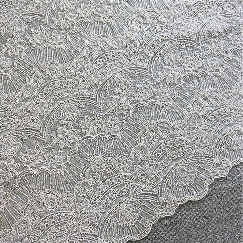 May include: White beaded lace fabric with a delicate floral pattern. The fabric features a repeating design of small, white beads arranged in a fan shape.