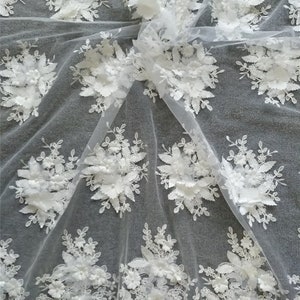 May include: White floral lace fabric with 3D flowers and leaves. The fabric is sheer and delicate, with a soft, flowing texture. The flowers are white and have a pearl-like finish. The leaves are green and have a realistic look.