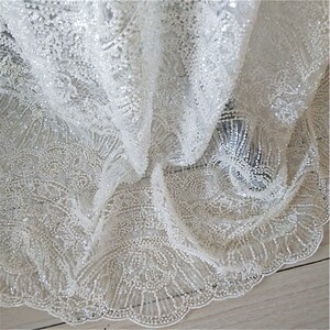 May include: White beaded lace fabric with a delicate floral pattern. The fabric is sheer and has a subtle shimmer.