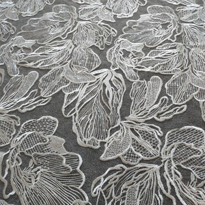 May include: White floral lace fabric with a delicate, intricate design. The lace features a repeating pattern of flowers with a textured, raised surface.