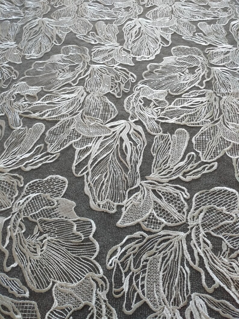 May include: White floral lace fabric with a delicate, intricate design. The lace features a repeating pattern of flowers with a textured, raised surface.